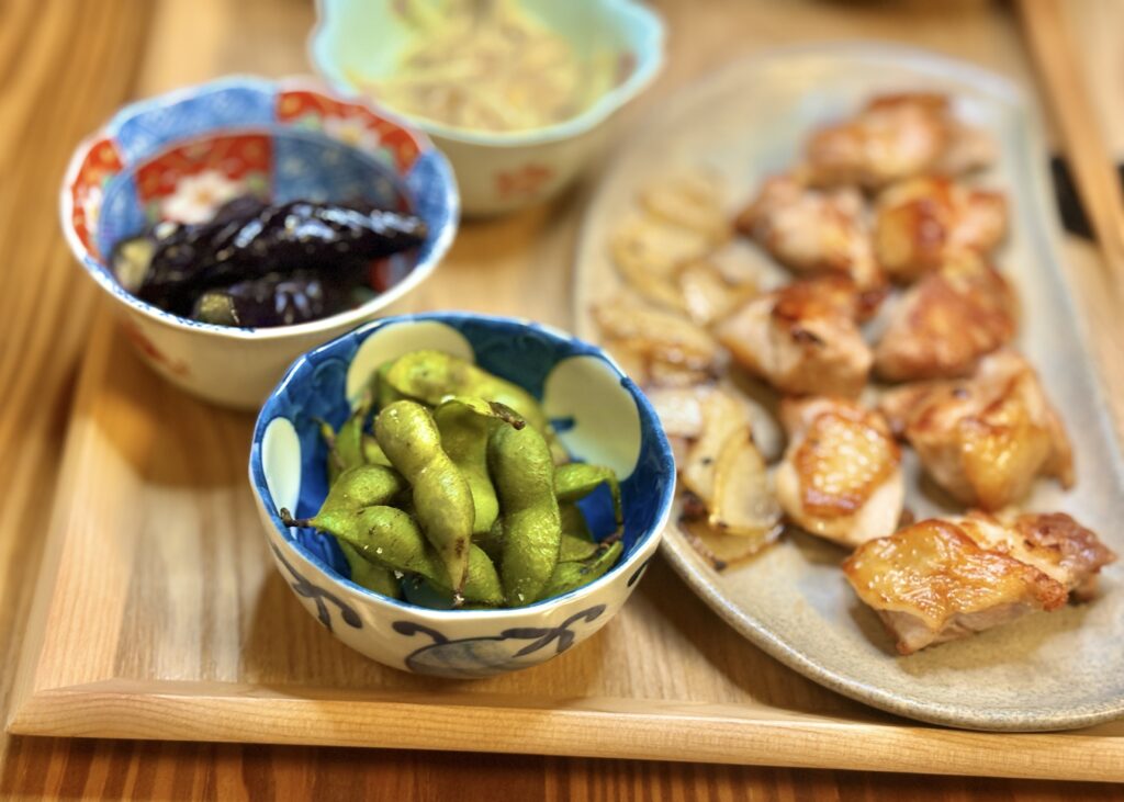 丹波篠山の黒枝豆とナスの煮浸しが並ぶ食卓。奥にはもやしのナムルと阿波尾鶏のグリルが見え、木のトレーの上に秋の味覚が彩りよく盛り付けられている