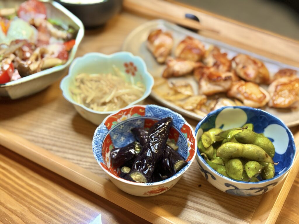 丹波篠山の黒枝豆とナスの煮浸し、もやしのナムルが並ぶ食卓。奥には阿波尾鶏のグリルと温玉シーザーサラダも見え、木のトレーの上に秋の味覚が彩りよく並んでいる。