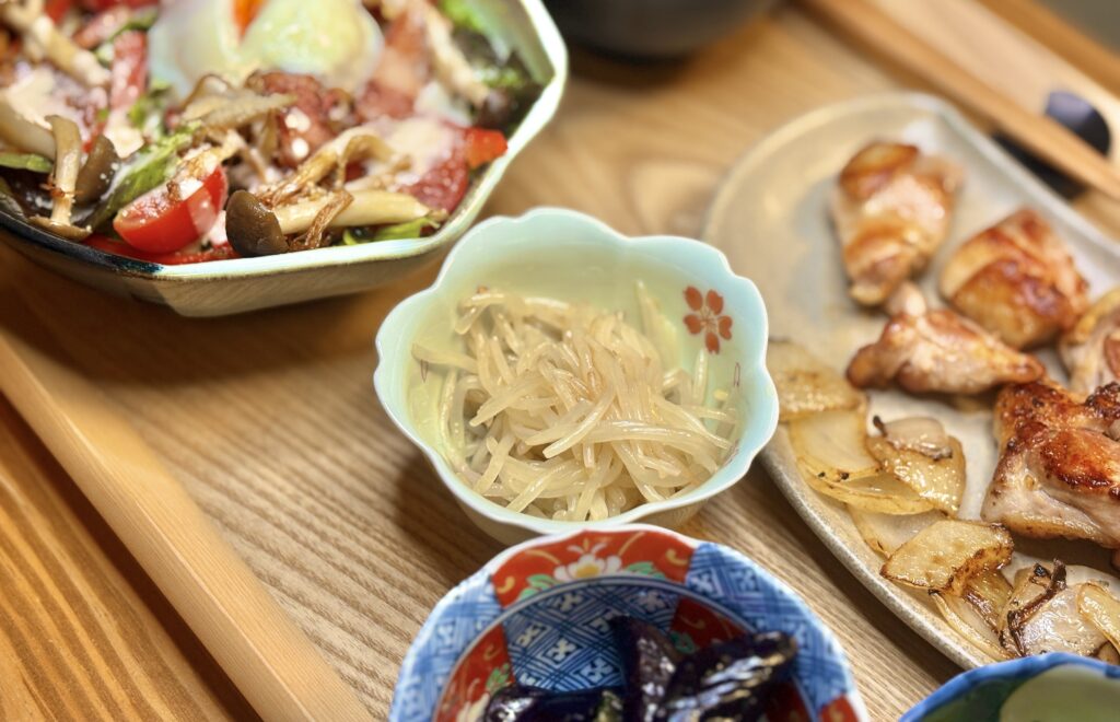 もやしのナムルを中心に、温玉シーザーサラダ、阿波尾鶏のグリル、ナスの煮浸しなどが並ぶ食卓。木目のトレーの上に、彩り豊かなおかずが丁寧に盛り付けられている。