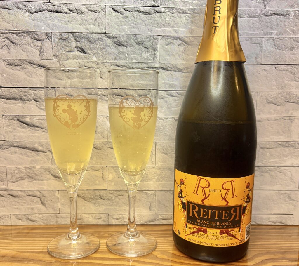 金色のラベルが印象的なスパークリングワイン『REITER ブラン・ド・ブラン』のボトルと、泡が立つシャンパングラス2つ。ミッキー＆ミニーの模様が入ったグラスが並び、華やかな乾杯のシーンを演出している。
