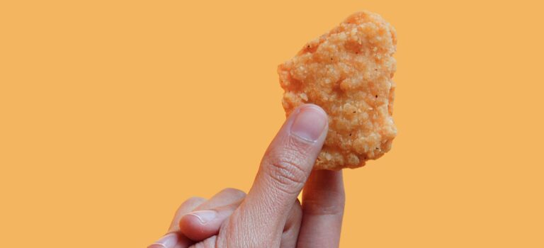 オレンジ色の背景に、手でつまんだ一口サイズのチキンナゲット。衣がサクッと揚がっており、シンプルでユーモラスな構図。