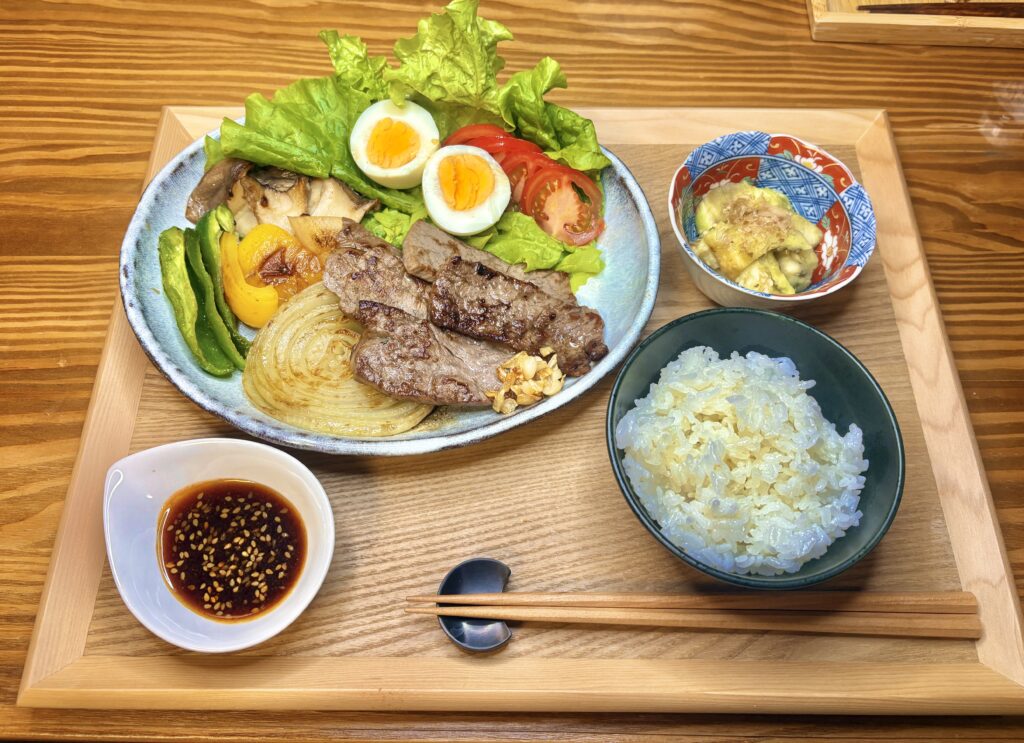木のトレイに盛りつけられた焼肉定食の食卓。
メインの皿には牛ステーキ、焼きタマネギ、ピーマン、パプリカ、エリンギ、ゆで卵、トマト、レタスなどが彩りよく並び、右上の小鉢には焼きなすのおひたし。
左下に「モランボン 焼肉の生だれ ジャン」が入った小皿、右下にはつやつやの白ごはん。
バランスの取れたおうちごはんプレート。