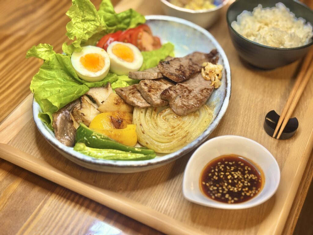 焼肉定食のおうちごはんプレート。
丸い皿に牛ステーキ、タマネギステーキ、エリンギ、ピーマン、パプリカ、レタス、トマト、半熟ゆで卵を盛りつけ、隣には炊きたての白ごはんと「モランボン 焼肉の生だれ ジャン」。
木のトレイに並んだ、彩り豊かでバランスの良い定食スタイルの食卓。
