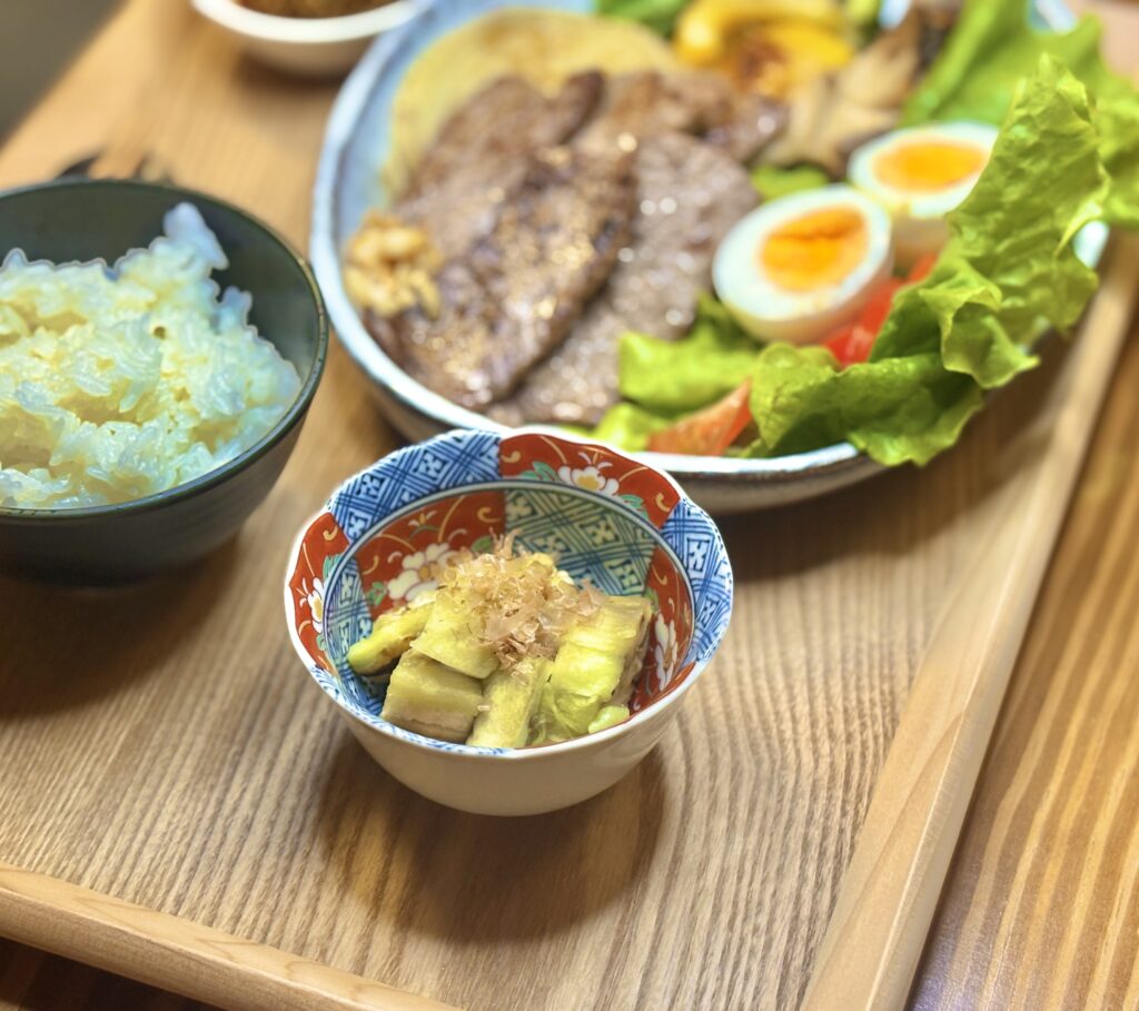 小鉢に盛りつけられた焼きなすの小皿料理。
彩り豊かな和柄の器に、熱々の焼きなすが盛られ、上にはかつお節がのっている。
背景には白ごはんと焼肉定食のプレートがぼんやり写り、温かみのあるおうちごはんの雰囲気を伝える写真。