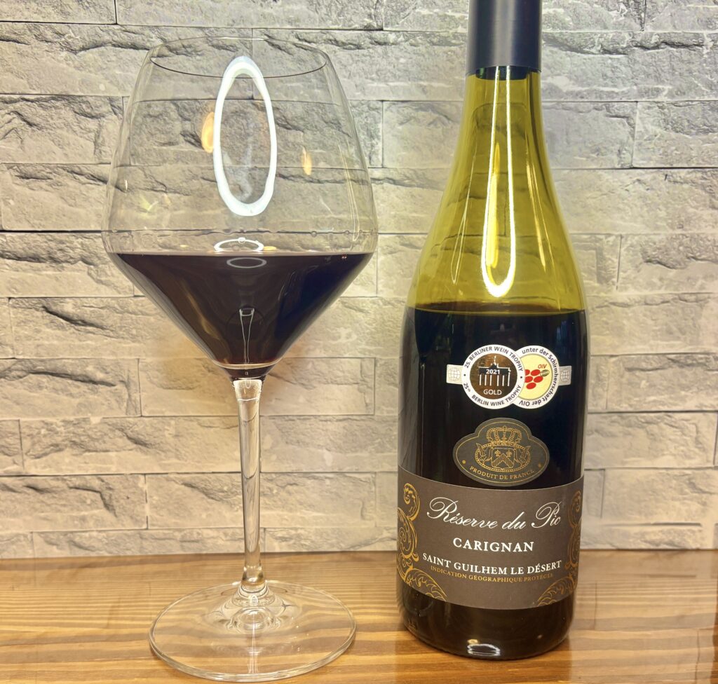 レゼルヴ・デュ・ピオ(カリニャン)の赤ワインボトルとグラスの写真。
深いルビー色のワインが入った大ぶりのグラスと、金のエンブレムが印象的なフランス産赤ワインのボトルが並んでいる。
木目のテーブルと白いレンガ調の背景で、落ち着いた雰囲気のワインシーンを演出。
