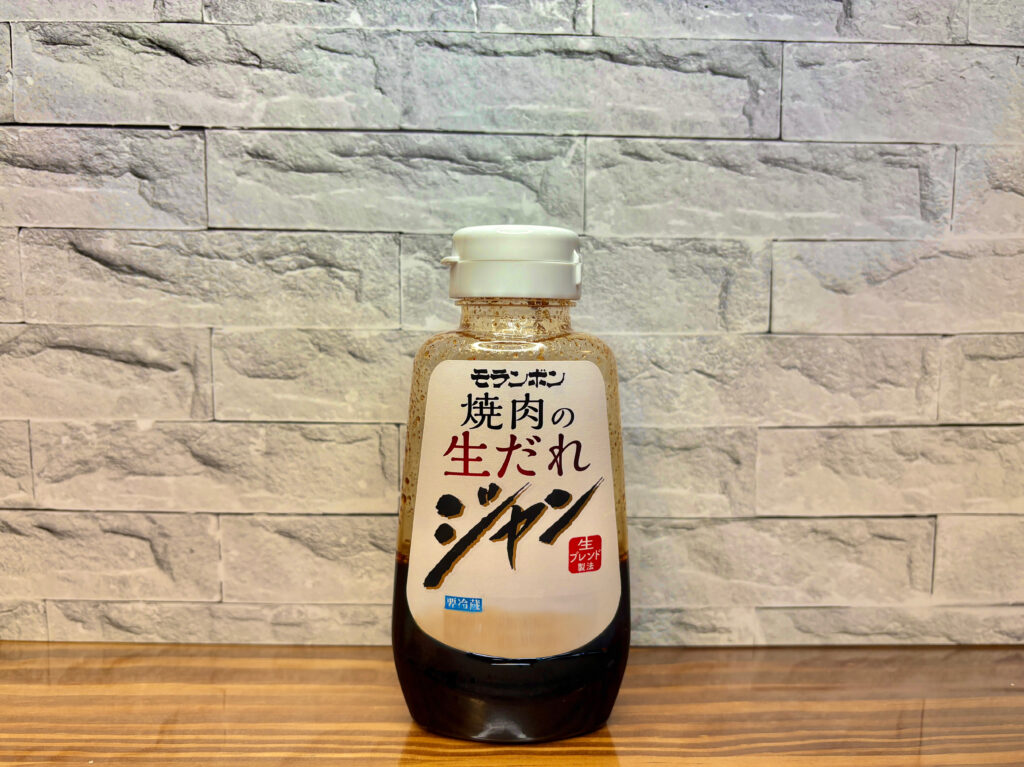 モランボン「焼肉の生だれ ジャン」のボトル写真。
白いラベルに黒文字で商品名が書かれ、赤い“生ブレンド製法”の印が目立つ。
木目のテーブルと白いレンガ調の背景で撮影された、焼肉定食に使われたタレのイメージ写真。