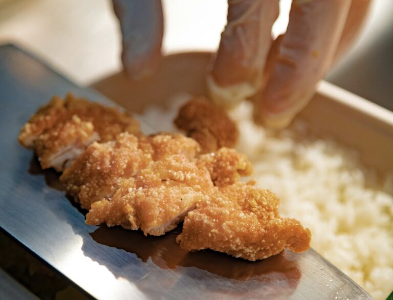 ご飯の上にサクサクに揚がったチキンカツを丁寧に盛りつける様子。 衣の香ばしさと照りが伝わる、できたての温かいお弁当シーン。