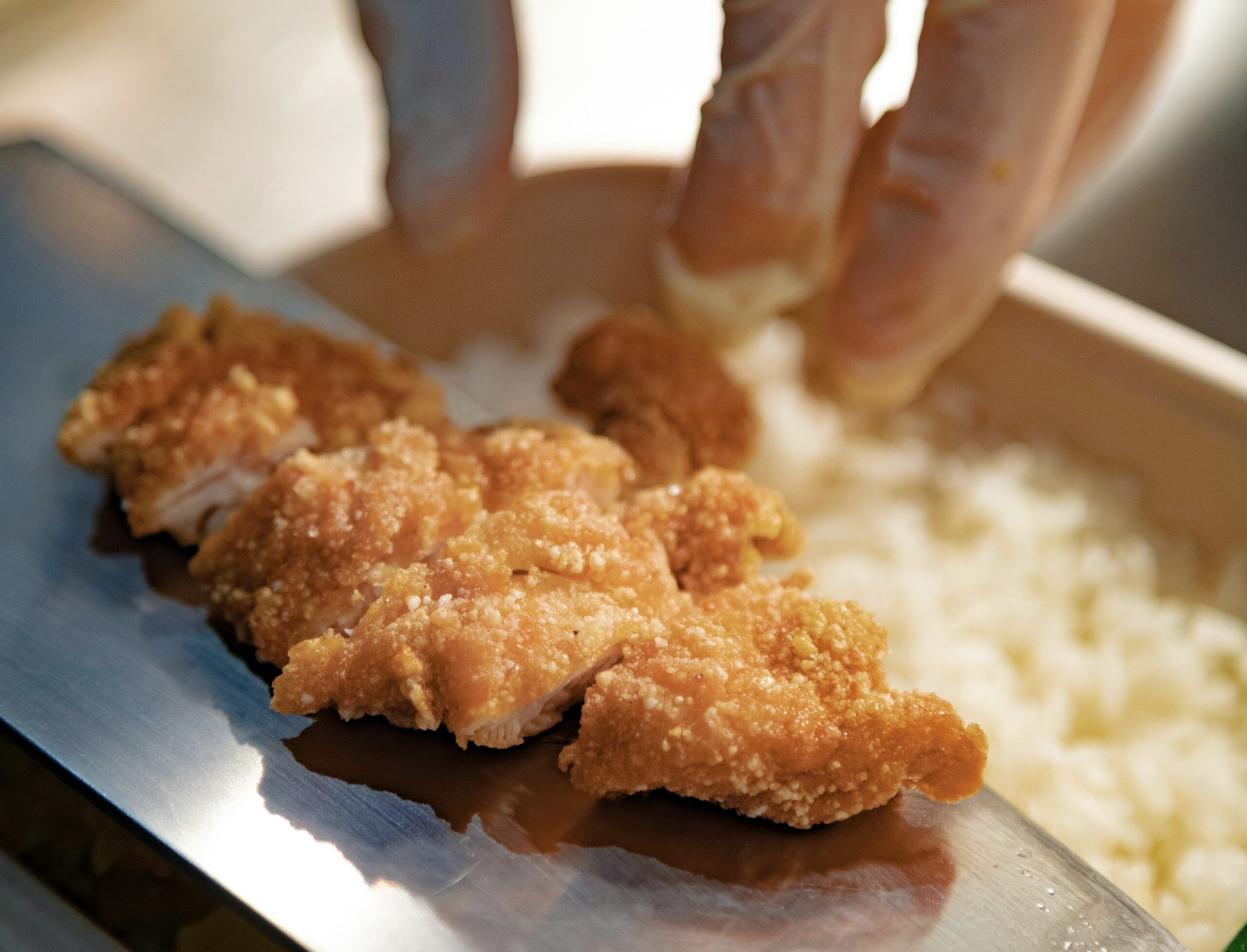 ご飯の上にサクサクに揚がったチキンカツを丁寧に盛りつける様子。 衣の香ばしさと照りが伝わる、できたての温かいお弁当シーン。