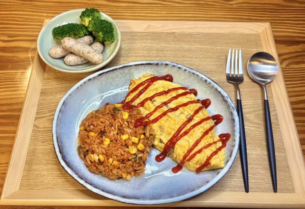 木のトレーに盛りつけられたチキンライス添えのじゃがいも入りオムレツ。
ケチャップがストライプ状にかかり、彩り豊かな家庭の夜ごはんプレート。
奥にはブロッコリーとゆかり入りソーセージの小鉢が添えられている。