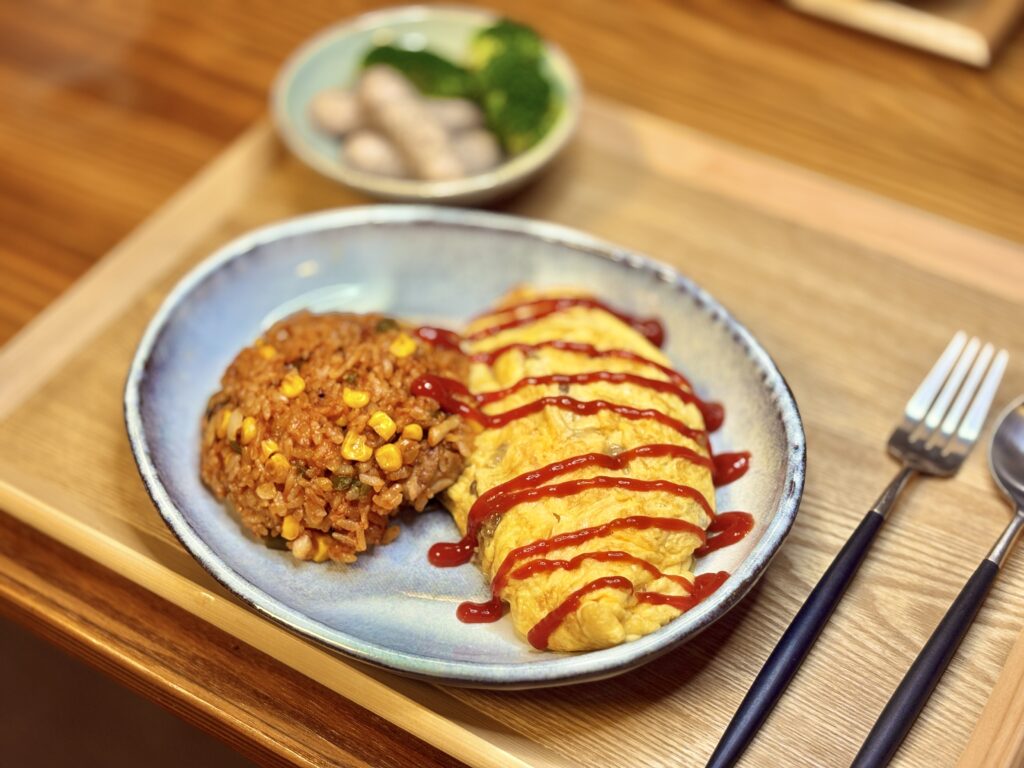 青みがかった楕円皿に盛られた、ふんわり焼かれたじゃがいも入りオムレツとチキンライス。
ケチャップがオムレツの上にストライプ状にかかり、彩り鮮やか。
奥にはブロッコリーとゆかり入りソーセージの小鉢が添えられた、木製トレーの上の家庭ディナー。