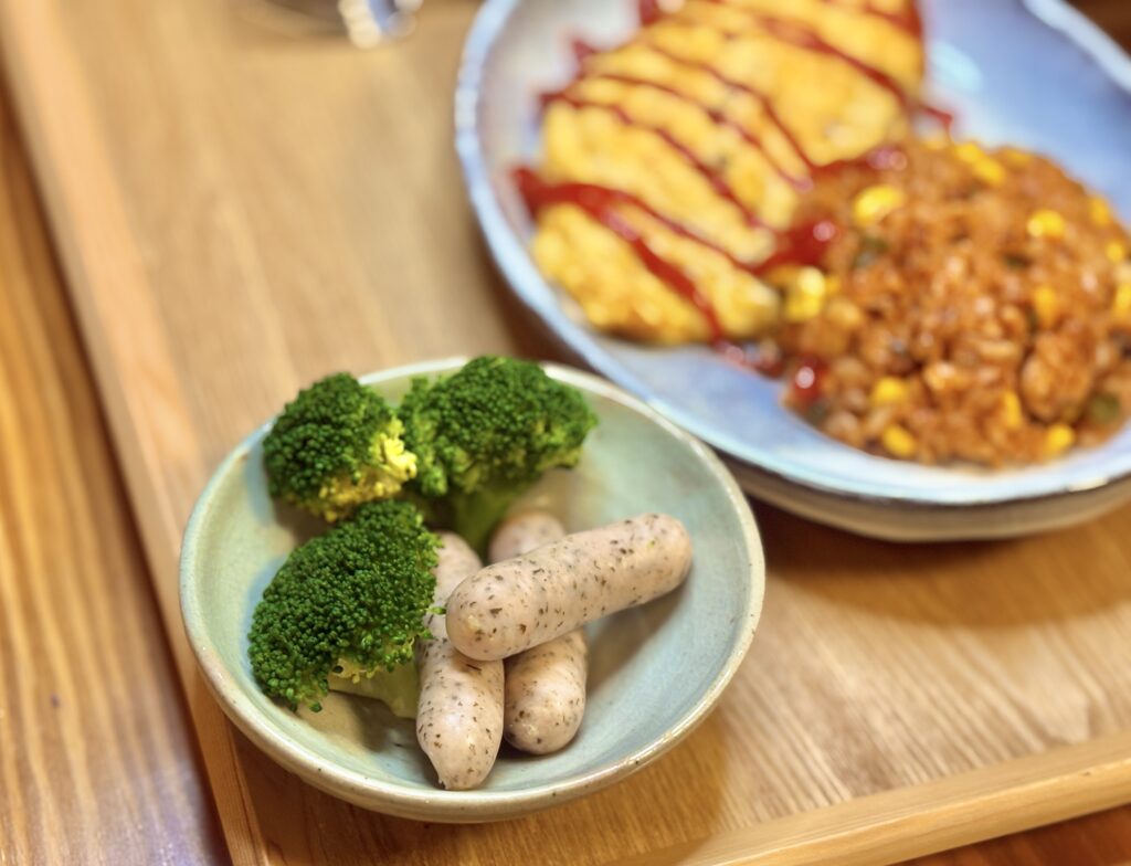 小さな器に盛りつけられた、ゆかり入りソーセージとブロッコリーの小鉢。
手前にはブロッコリーの鮮やかな緑、奥には粒ゆかりの入った白いソーセージが並び、
背景にはオムレツとチキンライスのプレートがぼんやり映る構図。