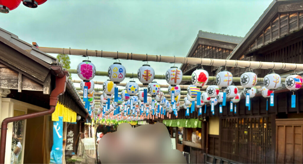 おかげ横丁の通りに色とりどりの提灯が並ぶ風景。和の雰囲気あふれる町並みに、観光客が行き交い、にぎやかで華やかな雰囲気が広がっている。