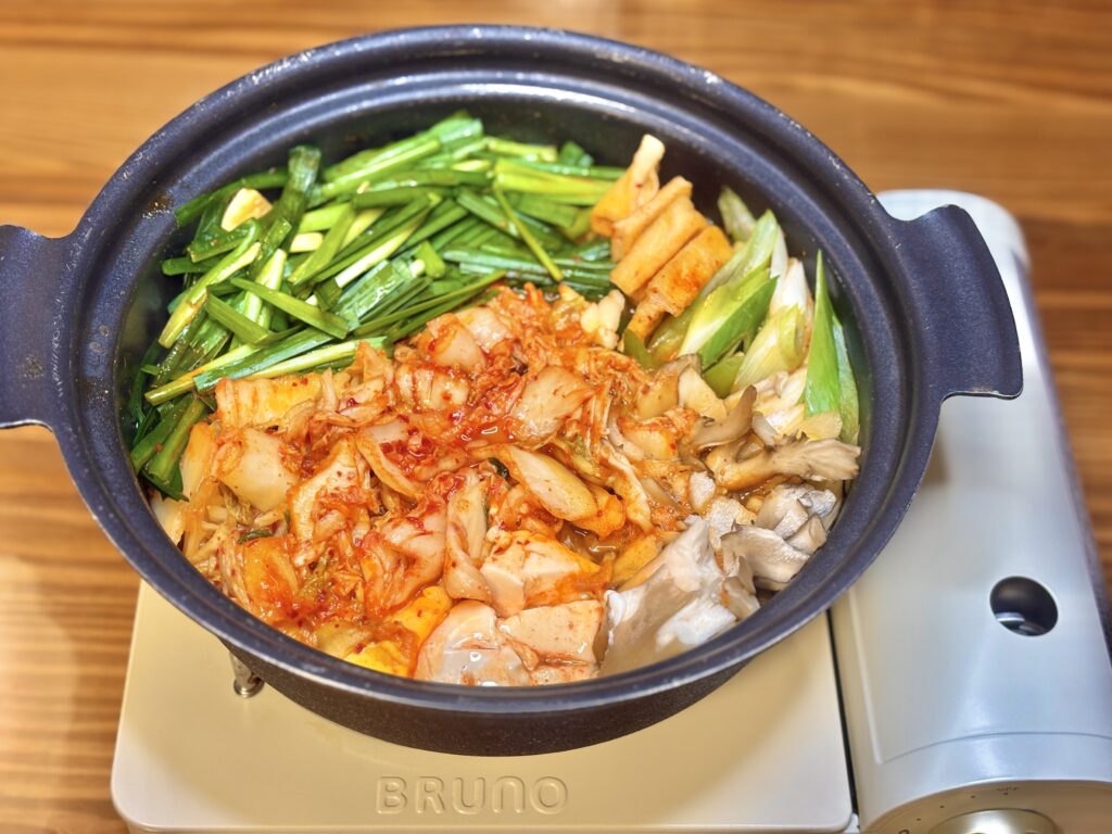 卓上コンロに置かれた黒い土鍋で、キムチ・ニラ・長ねぎ・まいたけ・厚揚げなど具材をたっぷり並べたキムチ鍋を上から撮影した写真