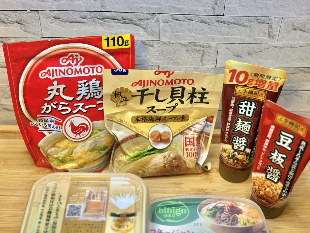 キムチ鍋に使う調味料を並べた写真。丸鶏がらスープ、干し貝柱スープ、甜麺醤、豆板醤などのパッケージがテーブルに立てて置かれている」
