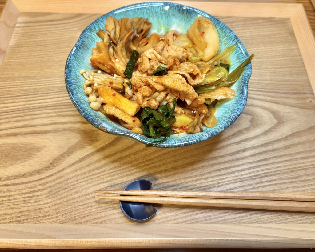 青い器に盛り付けたキムチ鍋の取り分け皿。豚肉、キムチ、まいたけ、えのき、厚揚げ、ニラなどの具材が彩りよく並び、横には箸が添えられている写真