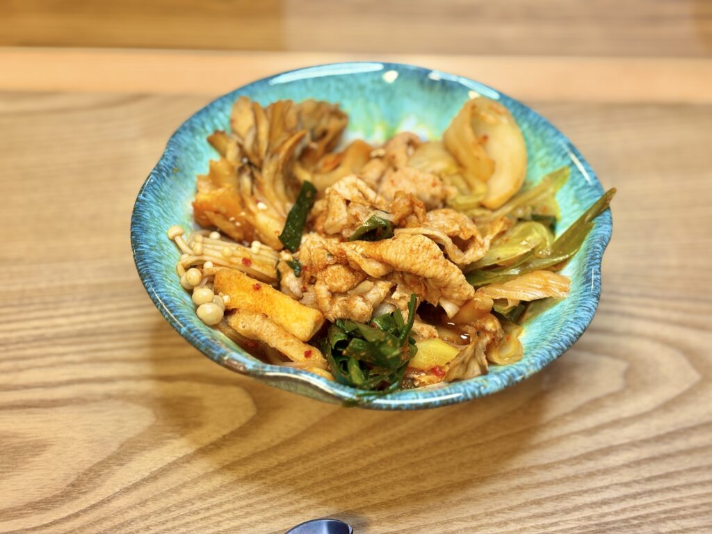 青い器に盛り付けたキムチ鍋の取り分け皿。豚肉、キムチ、まいたけ、厚揚げ、えのき、ニラ、長ねぎが彩りよく盛られた温かい一品のクローズアップ写真