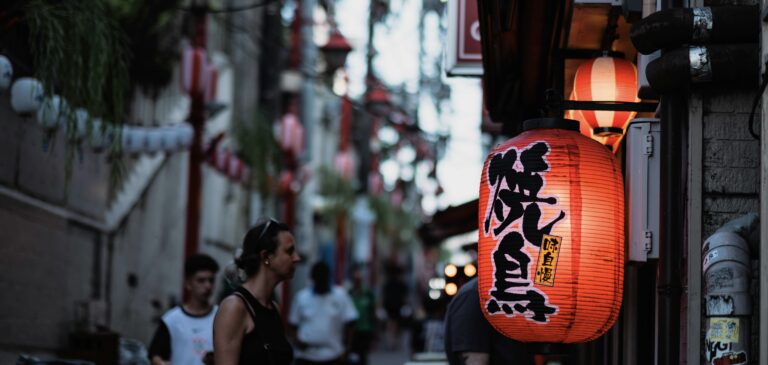 夕暮れ時の路地に灯る赤い提灯。大きく「焼鳥」と書かれた提灯が光り、奥には通りを歩く人々や並ぶ提灯の明かりがぼんやりと続く、ノスタルジックな街角の風景。