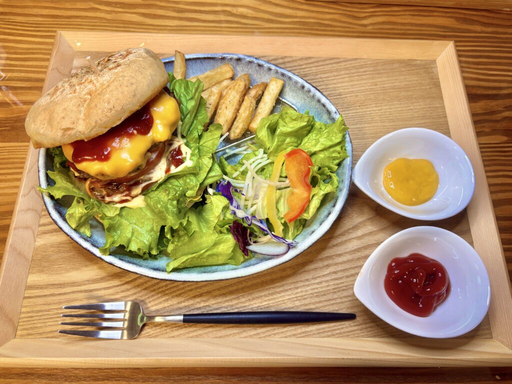 木のトレーに盛り付けられた、チーズたっぷりの手作りハンバーガーとポテト、彩り豊かなサラダ。横にはケチャップとマスタードが小皿に添えられている。