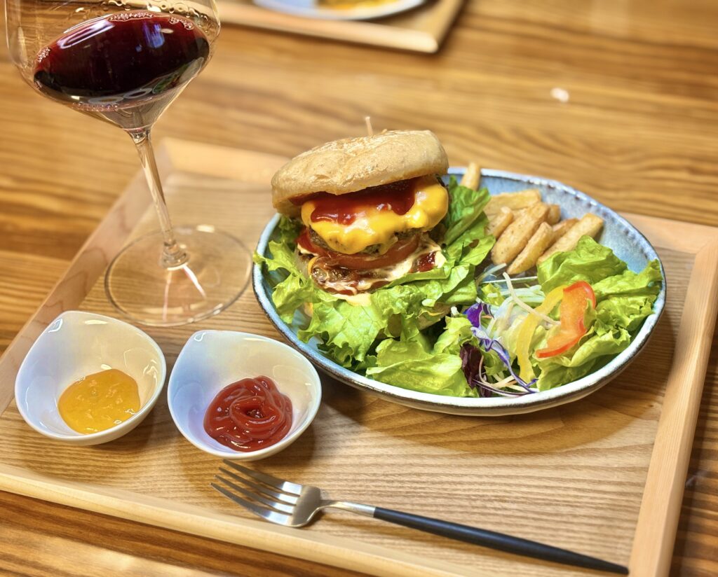 木のトレーの上に、チーズたっぷりの手作りハンバーガー、ポテト、彩り豊かなサラダが盛られたワンプレートと、横に赤ワインの入ったグラスが置かれている写真。
