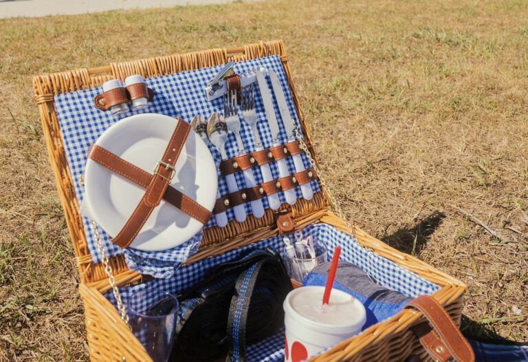 青と白のギンガムチェック柄の布を敷いたピクニックバスケット。皿やカトラリーが革ベルトで固定され、芝生の上に置かれている。手前にはドリンクカップが入っており、穏やかなピクニックの雰囲気が漂う。