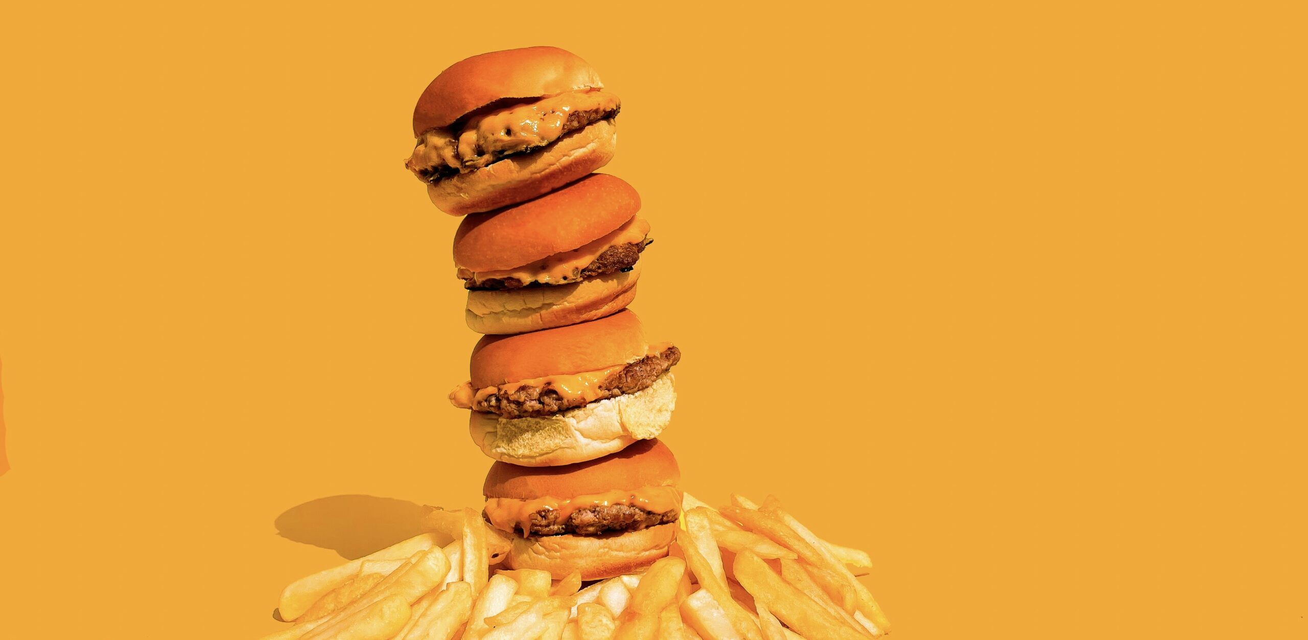 鮮やかなオレンジ色の背景に、チーズバーガーが縦に積み上がったタワーのように並び、足元にはたっぷりのフライドポテトが広がっている写真。