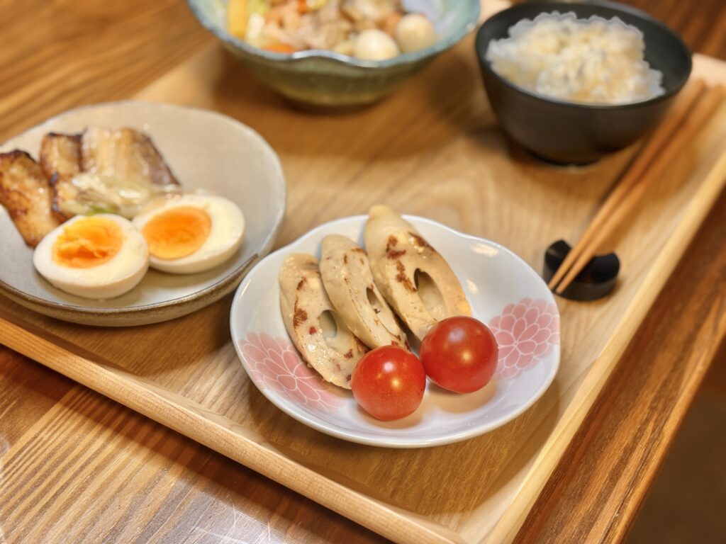豆腐ちくわとミニトマトの皿を中心に、ネギチャーシュー煮卵や白ごはん、八宝菜が木のトレイに並ぶ食卓の写真