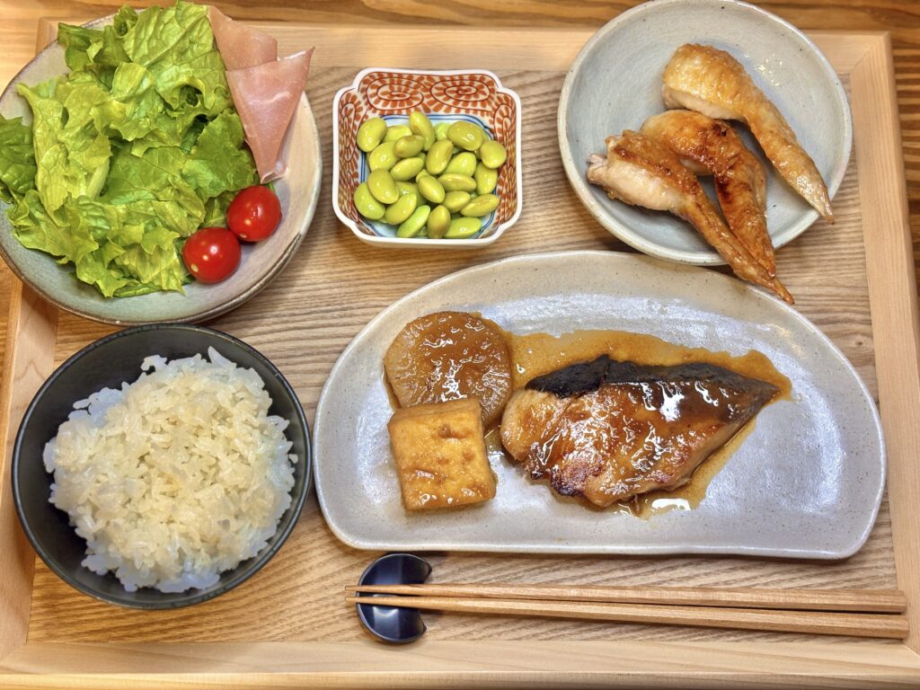 鰤の照り焼き、大根と厚揚げの煮物、手羽先の塩焼き、秘伝豆、生ハムサラダ、白ごはんを木のトレイに盛りつけた和食の定食。