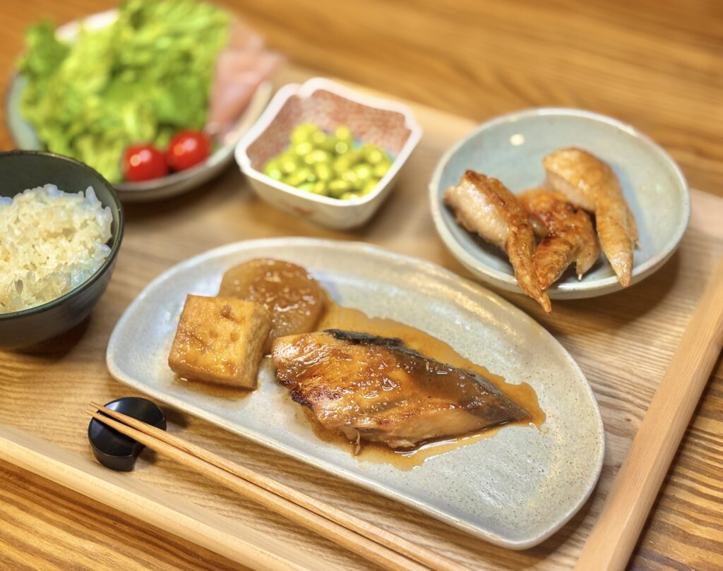 鰤の照り焼きを中心に、手羽先の塩焼き、秘伝豆、生ハムサラダ、白ごはんを並べた和食の食卓の写真。