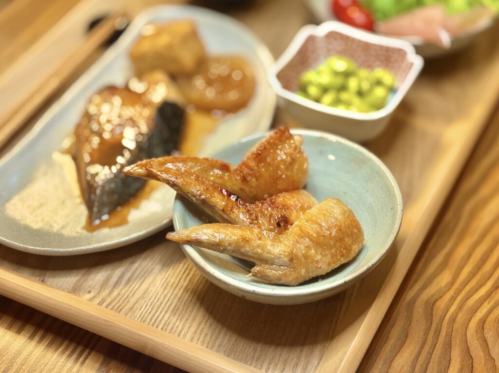 こんがり焼き上げた手羽先の塩焼きを小皿に盛りつけたアップ写真。奥には鰤の照り焼きや秘伝豆が見える。