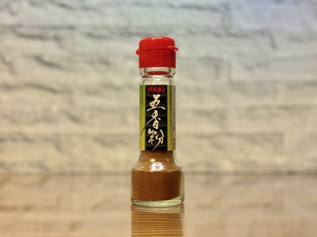 ユウキ食品の五香粉（ウーシャンフェン）の瓶がテーブルに置かれている。赤いキャップと黒いラベルが特徴で、中華料理のアクセントに使われるスパイスが入っている。