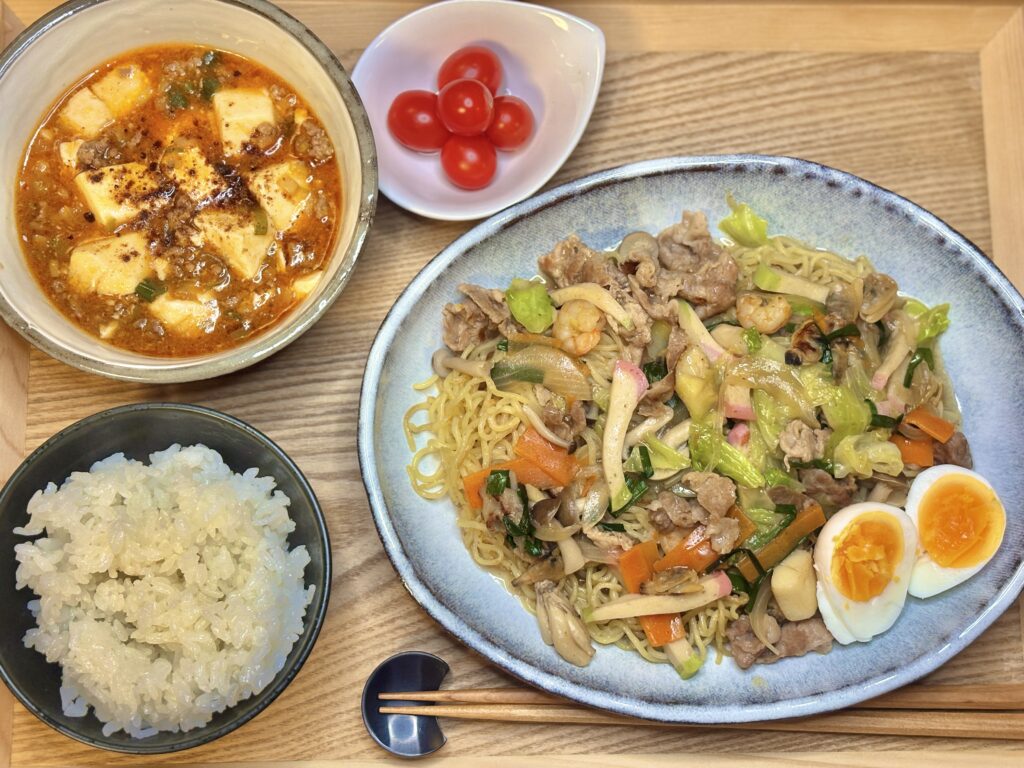 木製トレーの上に並べられた中華風の晩ごはんセット。
具だくさんの餡掛け焼きそば、半熟ゆで卵、コクのある麻婆豆腐、白ごはん、彩りのミニトマトがきれいに配置されている。温かみのある食卓で、家庭料理の豊かさが伝わる。