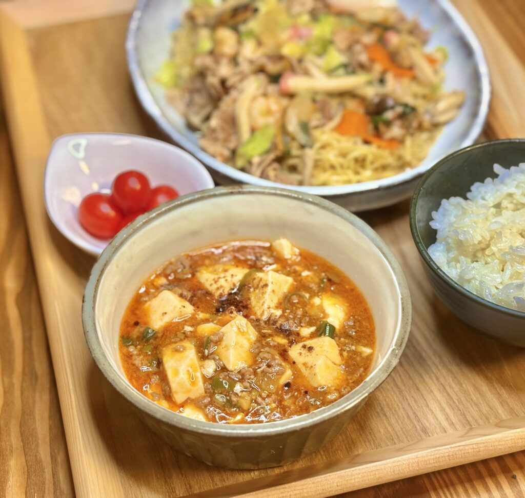 赤みのあるタレに大きめに切られた豆腐とひき肉がたっぷり入った麻婆豆腐のクローズアップ。奥に餡掛け焼きそばやごはん、ミニトマトがぼんやり写っている。