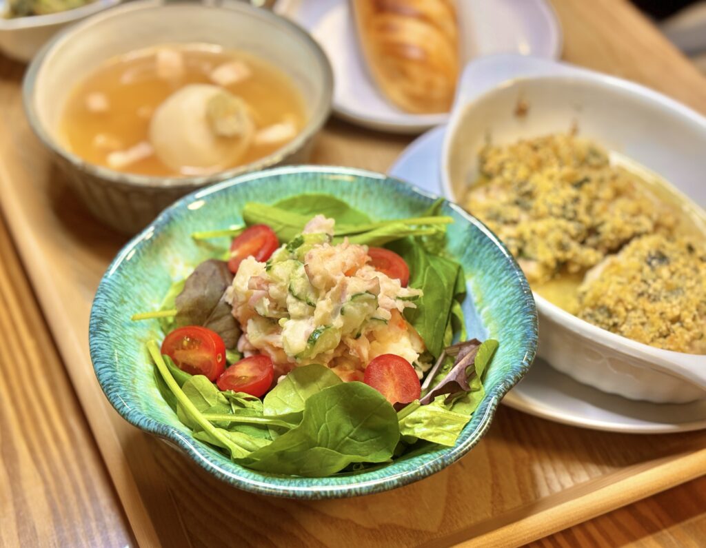 色鮮やかなベビーリーフとポテトサラダを主役に、香草パン粉焼きやスープが奥に並ぶ食卓の写真。