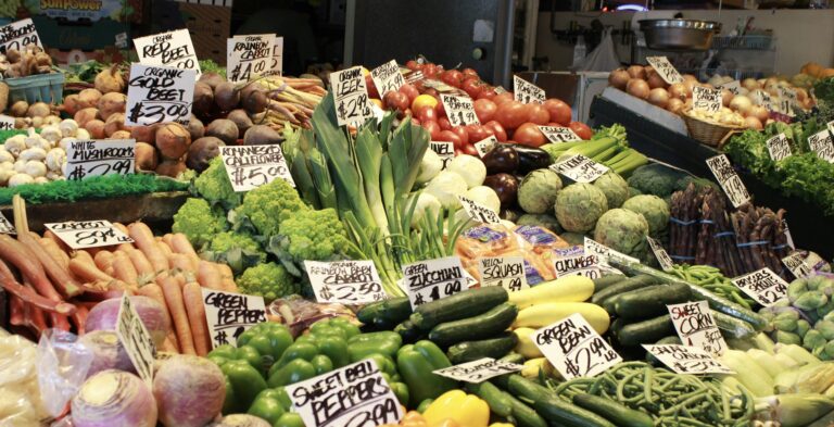 市場の野菜売り場に色とりどりの新鮮な野菜が並んでいる様子。 にんじん、ズッキーニ、トマト、きのこ、アスパラガス、ねぎ、アーティチョークなどが山盛りに陳列され、手書きの価格札が添えられている。 彩り豊かで活気あるマーケットの雰囲気が伝わる。