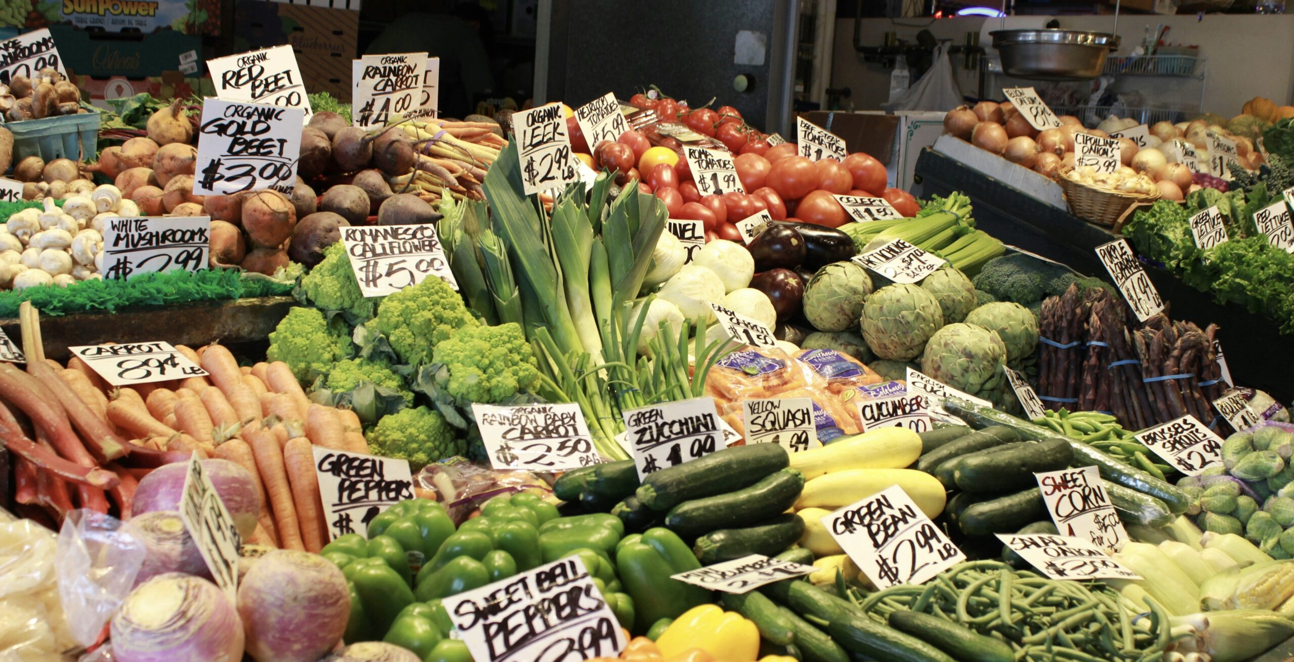 市場の野菜売り場に色とりどりの新鮮な野菜が並んでいる様子。 にんじん、ズッキーニ、トマト、きのこ、アスパラガス、ねぎ、アーティチョークなどが山盛りに陳列され、手書きの価格札が添えられている。 彩り豊かで活気あるマーケットの雰囲気が伝わる。