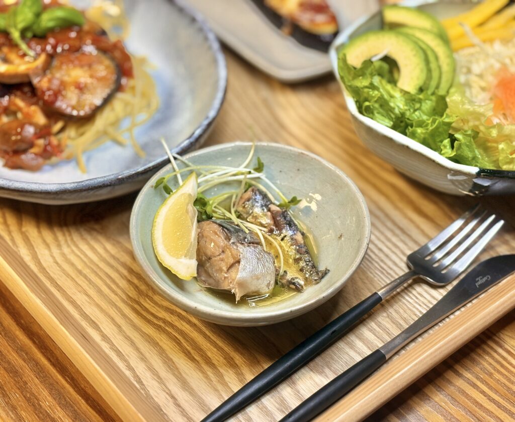 レモンを添えたオイルいわしの小鉢と、奥にパスタとサラダが見える食卓ショット
