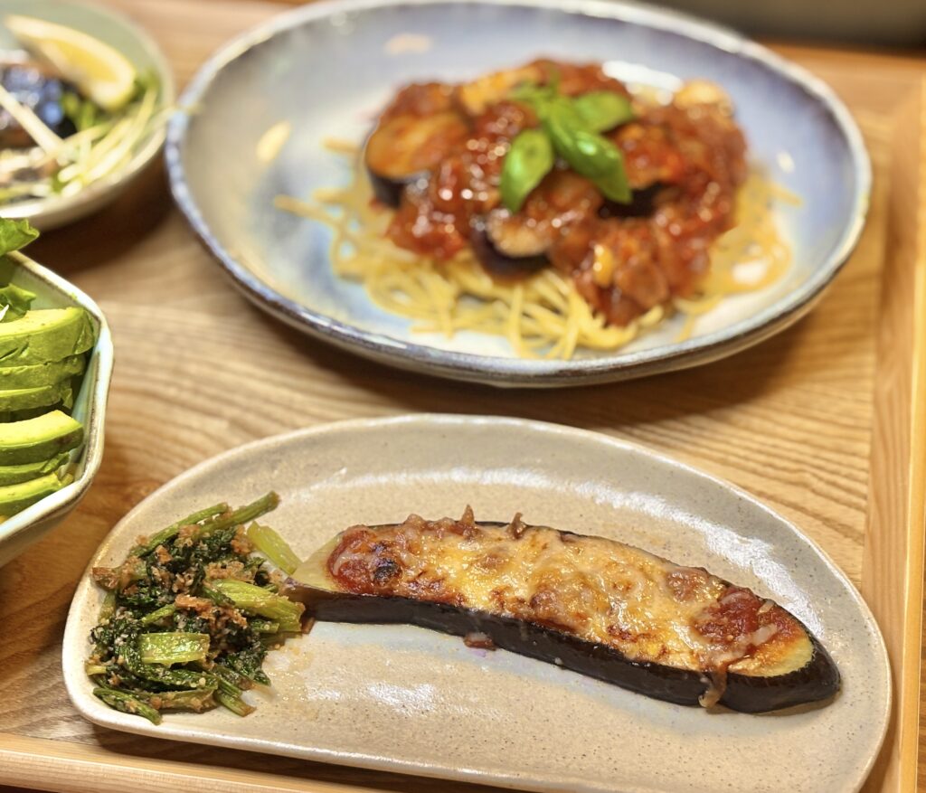 チーズをのせて焼いたナスのピザ風とほうれん草のおかか和え、奥には具だくさんトマトパスタが見える食卓ショット