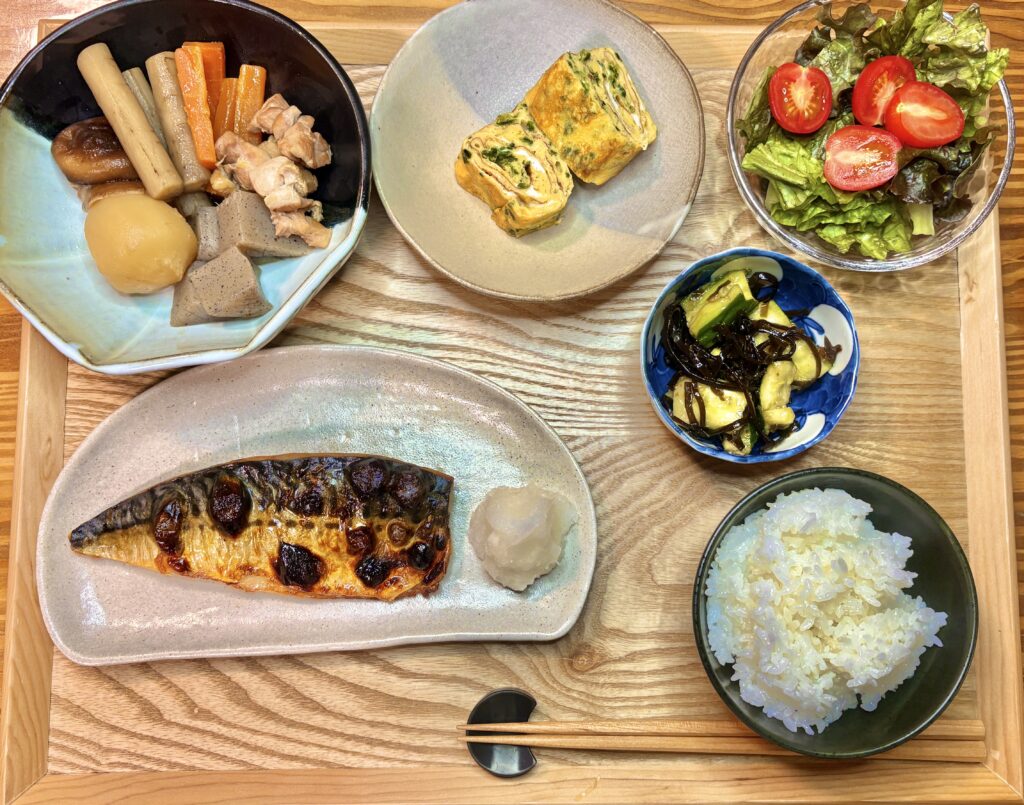 木のトレーに彩りよく並んだ家庭の和食献立。焼きサバ、白ごはん、だし巻き卵、根菜の煮物、トマト入りグリーンサラダ、モズク酢ときゅうりの小鉢が並び、全体が明るく健康的な食卓の上から写真