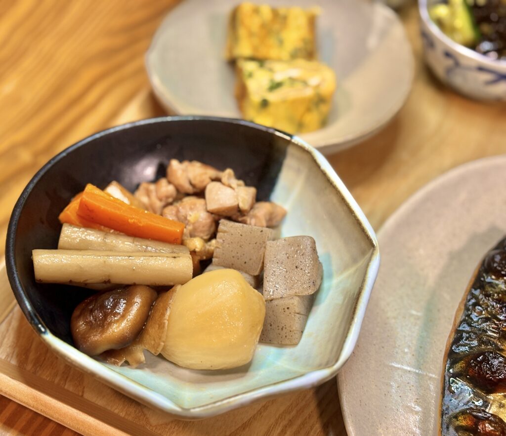 黒い器に盛られた根菜の煮物。にんじん、ごぼう、しいたけ、こんにゃく、鶏肉、そしてつやのあるじゃがいもが、優しい色合いで並ぶ。木目のテーブルの上で、家庭的で温かい雰囲気の一皿
