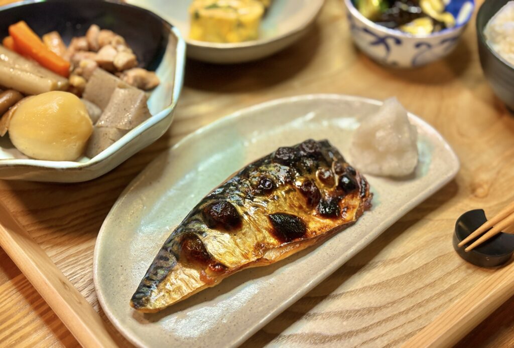 木目のテーブルに置かれた長皿に、こんがり焼けた塩サバが盛り付けられている。皮目は照りと焦げ目が混ざり、横に添えられた大根おろしがふんわりと白く映える。和食らしい落ち着いた雰囲気の一皿を写した写真