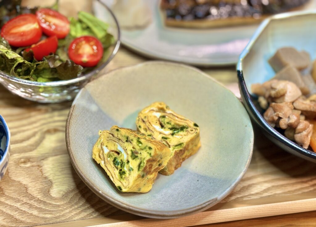 木のトレーの上、淡い色の丸皿にほうれん草入りのだし巻き卵が二切れ並んでいる。断面の渦巻き模様がふっくらと見え、黄色と緑のコントラストが鮮やかに映る和食の一品