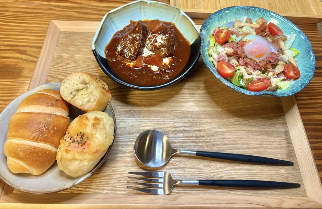 「ごろっと牛肉のビーフシチュー、温玉シーザーサラダ、カンテグランデの塩パン・明太チーズパン・ガーリックフランスを並べたディナーセット。木製トレーに映えるおうちレストラン風の食卓」