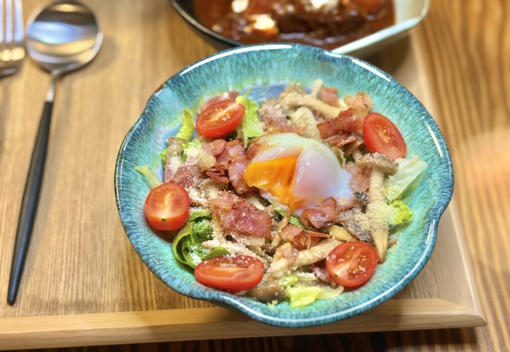 「温泉卵をのせたシーザーサラダ。とろりとした黄身がレタスやベーコン、プチトマトに絡む、おうちレストラン風の彩りサラダ」