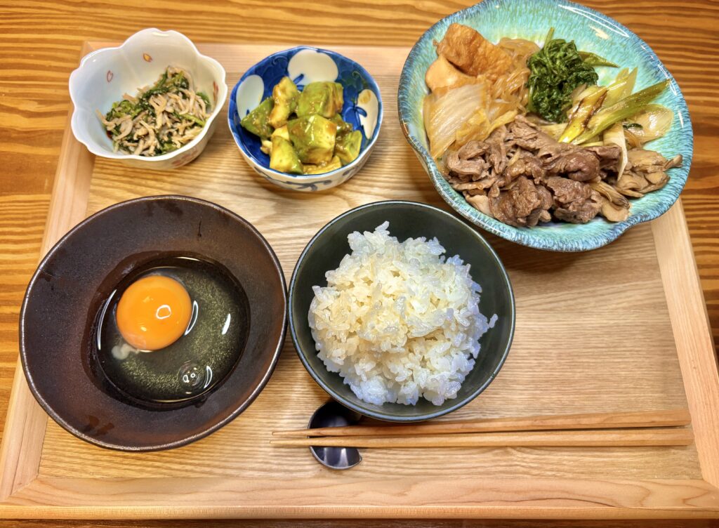 牛肉と野菜のすき煮、アボカドのわさび醤油和え、大根葉としらすのごま油炒めを盛り合わせた和食定食。生卵と白ごはんを添えた旦那キッチンの晩ごはん