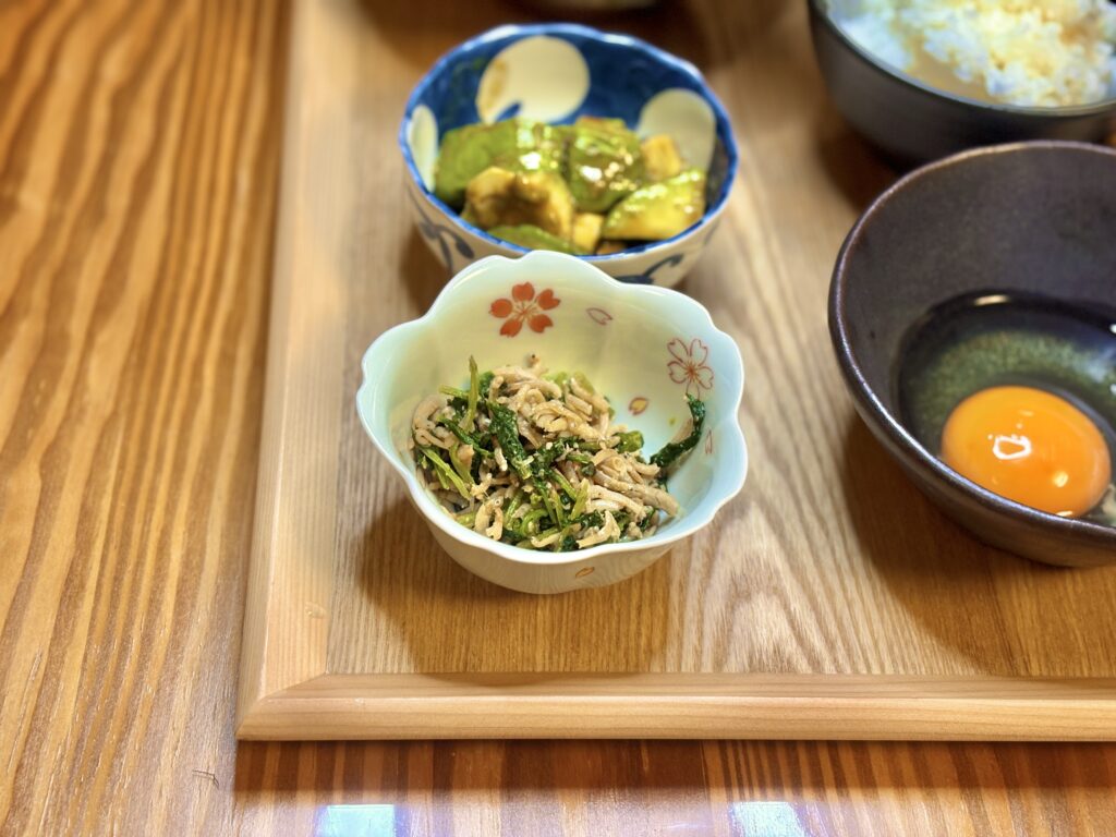 大根葉としらすをごま油で炒めた小鉢。香ばしい香りと塩気がご飯に合う一品。奥にはアボカドのわさび醤油和えと生卵が並ぶ