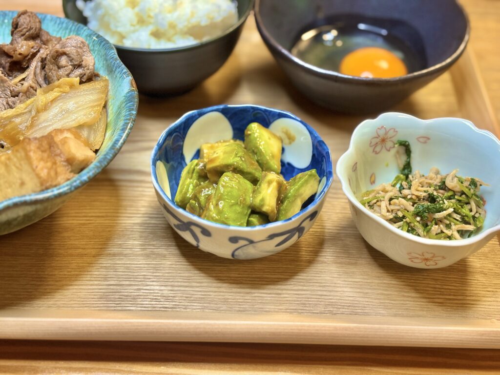 アボカドのわさび醤油和えと、大根葉としらすのごま油炒めの小鉢。奥にはすき煮とご飯、生卵が並ぶ旦那キッチンの和食プレート