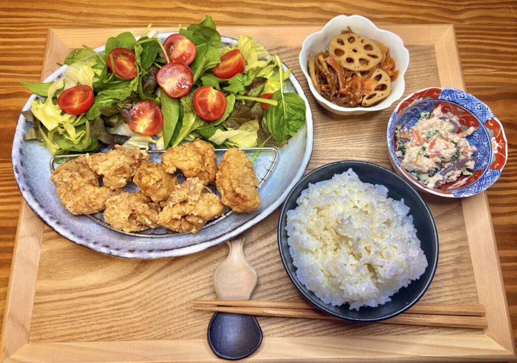唐揚げ、れんこんのきんぴら、白和え、たっぷりのサラダと白ごはんを並べた和定食。木のトレイに彩りよく盛り付けられた、バランスのとれた夕食。