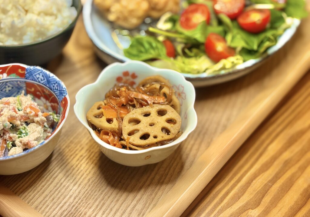 れんこんとにんじんのきんぴら。甘辛い照りが食欲をそそる家庭の副菜で、彩りも豊か。木のトレイの上に白和えやサラダと並ぶ温かみのある食卓の一皿。