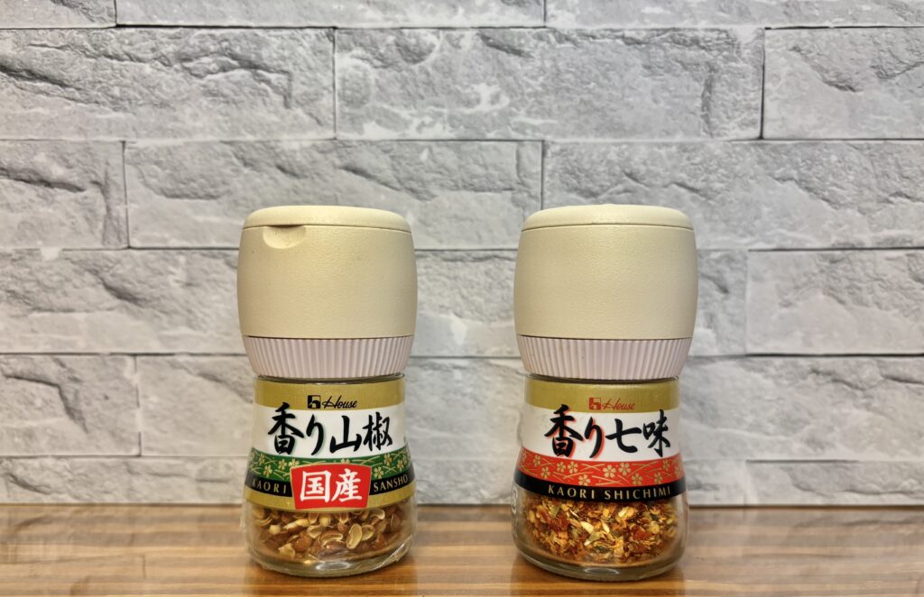 ハウスの「香り山椒」と「香り七味」。挽きたての香りが楽しめるミル付きスパイスで、料理の仕上げにぴったり。木目のテーブルの上に並べられた2本の調味料ボトル。