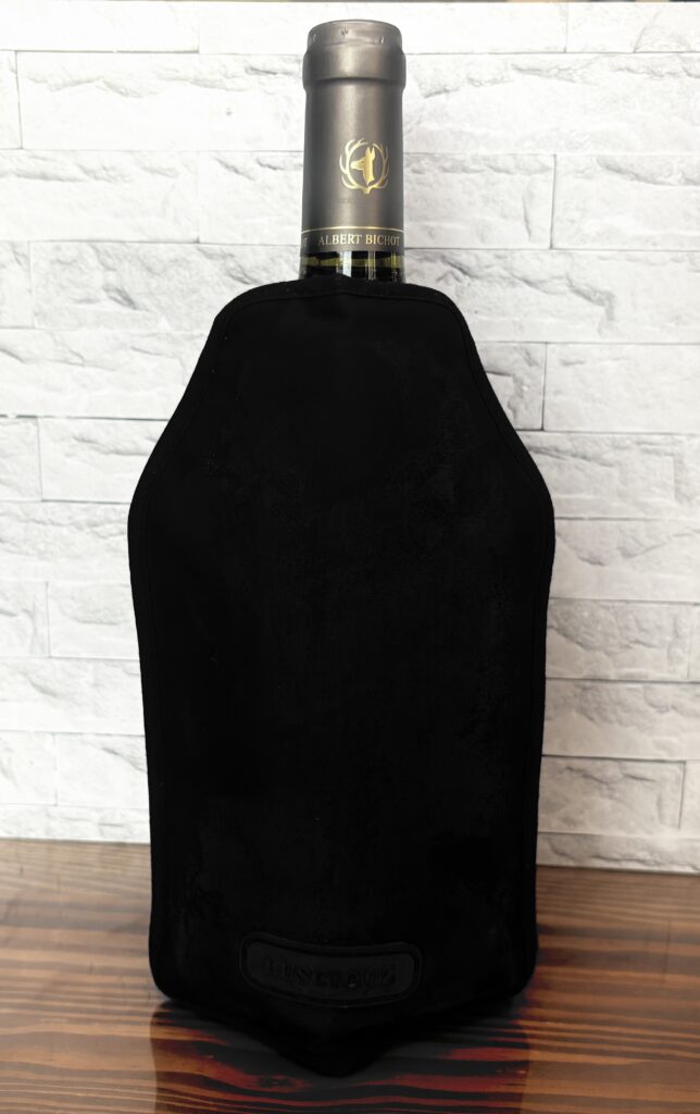 黒いボトルクーラーを装着した白ワインボトル。テーブルの上に立てられ、背後には白いレンガ調の壁。冷たさをキープしながら、見た目もスマートな印象のワインクーラー。